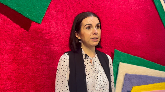 A woman with shoulder-length dark hair is standing in front of a vibrant red background featuring abstract green and white shapes. She is wearing a polka-dot blouse with a black vest and small hoop earrings, speaking with an expressive demeanor. A microphone is clipped to her vest.