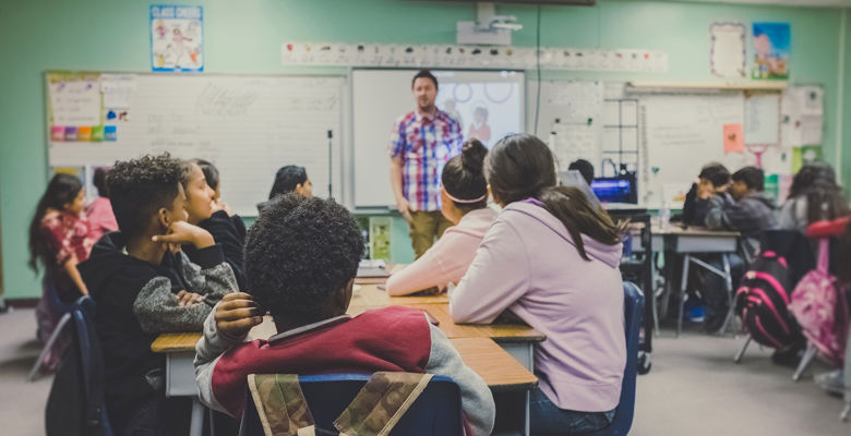 Classroom of young students looking towards their teacher. Several white boards are in the background.