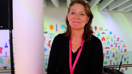 A woman with shoulder-length light brown hair is standing indoors, smiling warmly. She is wearing a black top, a pink lanyard, pearl earrings, and a pearl necklace layered with another pendant. Behind her is a white pillar, and the wall features colorful, abstract shapes and patterns under a modern ceiling structure. A microphone is clipped to her outfit.