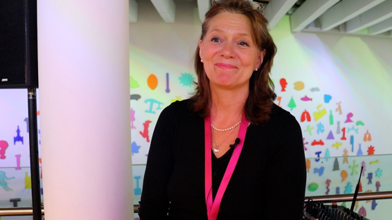 A woman with shoulder-length light brown hair is standing indoors, smiling warmly. She is wearing a black top, a pink lanyard, pearl earrings, and a pearl necklace layered with another pendant. Behind her is a white pillar, and the wall features colorful, abstract shapes and patterns under a modern ceiling structure. A microphone is clipped to her outfit.