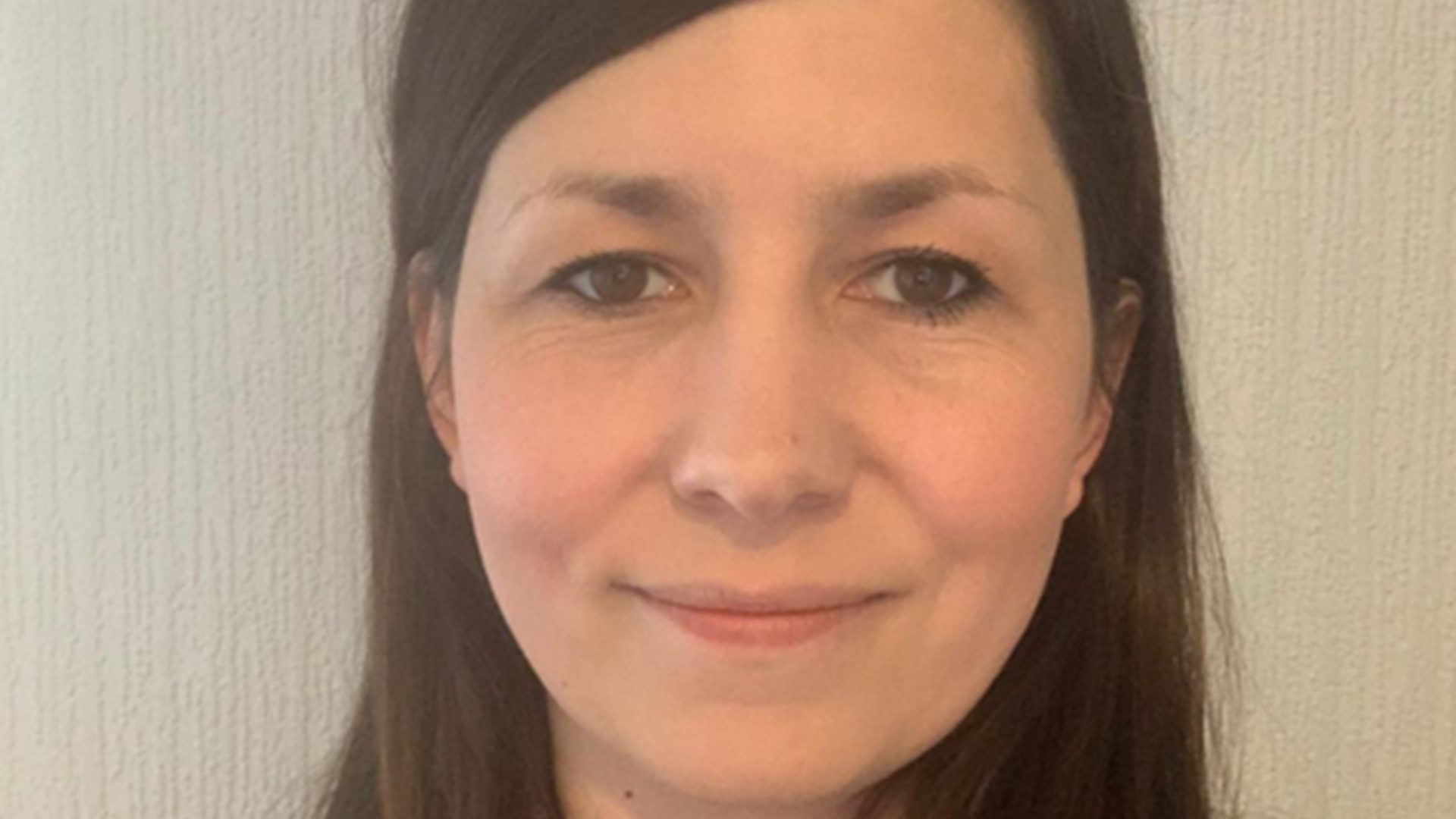 A person with long, straight brown hair standing in front of a textured, light‑coloured wall.