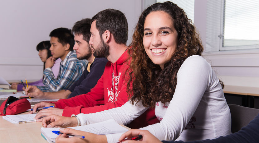 Smiling student is sitting at a desk with their peers, all holding pens and with notebooks in front of them.