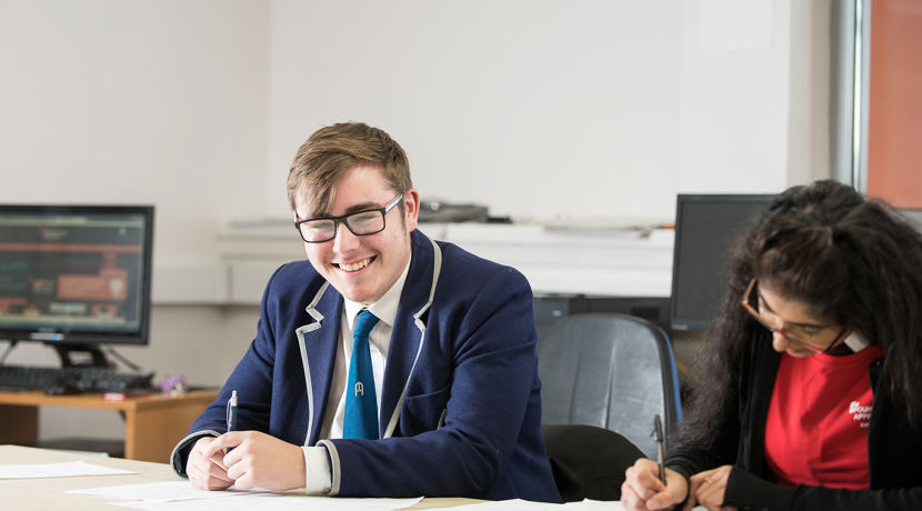 Young boy in a blue school uniform blazer and shirt and tie looks directly at the camera and smiles. He holds a pen in his hand. A young female is sitting next to him writing in her notepad.