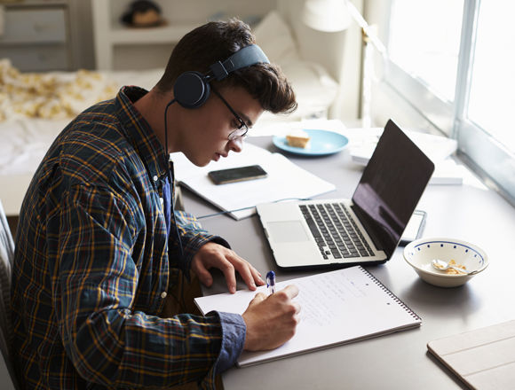 Young student wearing headphones, writing in a notebook with their laptop open on the desk.