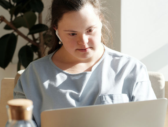 Student with earphones looking at their laptop with a large plant situated behind them.