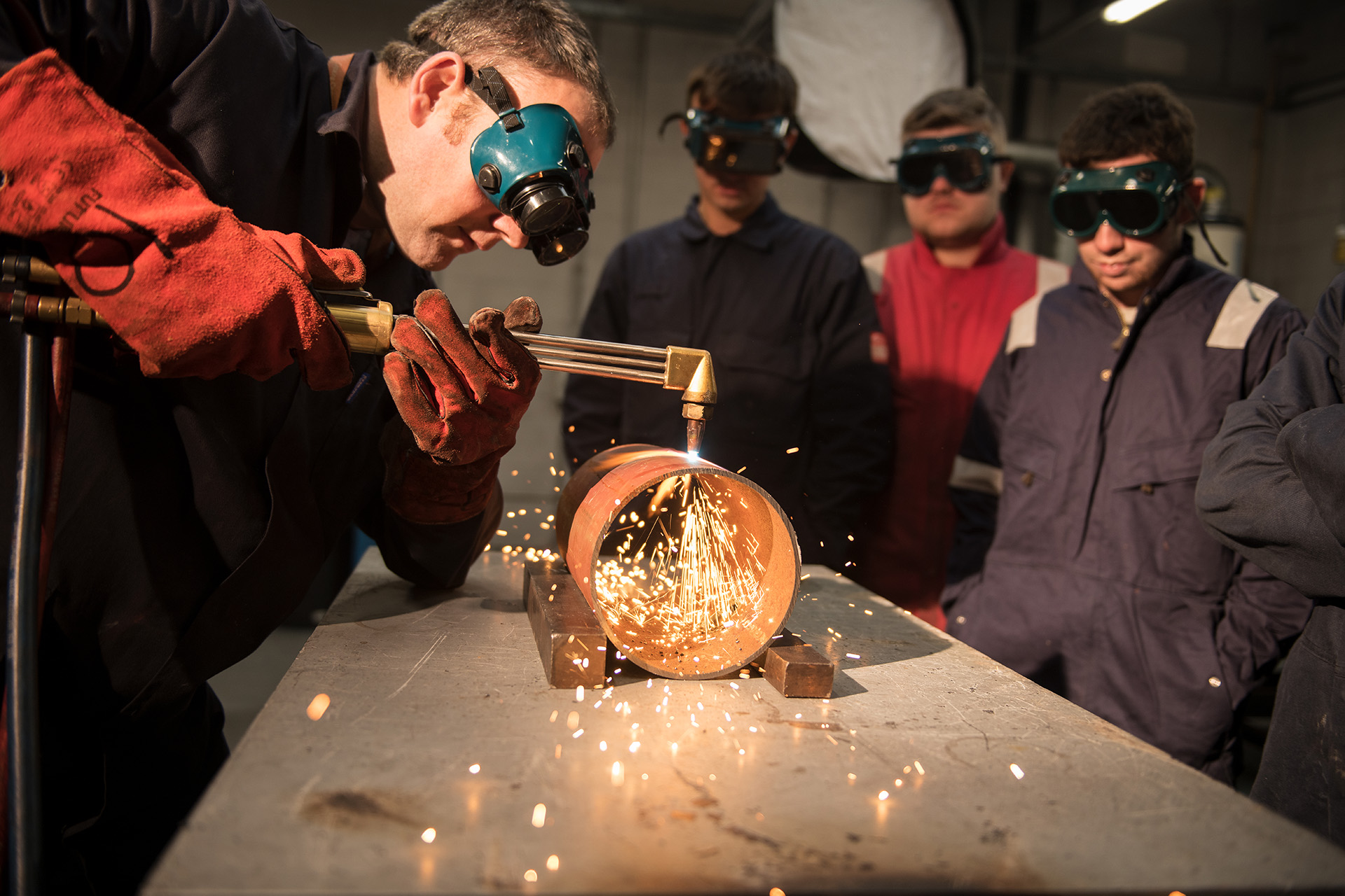 Engineering student wearing googles and a boiler suit is welding in front of peers, sparks are flying.
