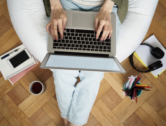 Shot from above, a person is sitting on a bean bag and typing on a laptop. The bean bag is surrounded by stationery, notebooks and a cup of cofee.