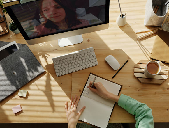Person writing notes at their desk with their computer on and a cup of coffee next to them.
