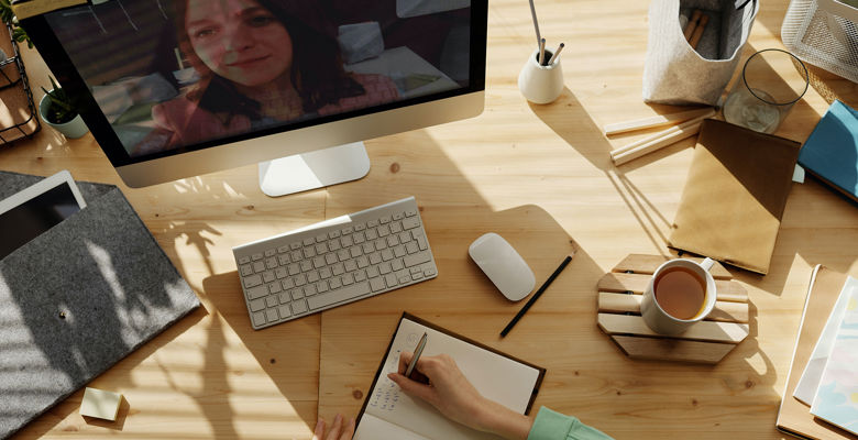 Person writing notes at their desk with their computer on and a cup of coffee next to them.