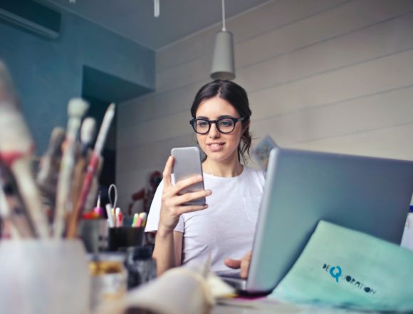 Young woman working at a cluttered desk with their laptop open and looking at their smartphone.
