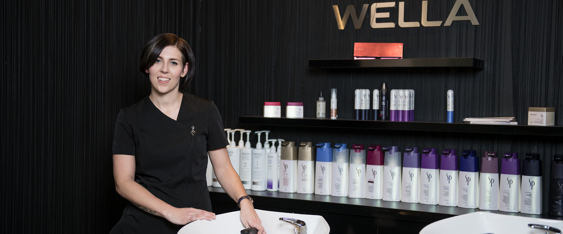 Hairdressing student smiling while they stand next to the shampoo section of the salon, with chair basins and products next to them.