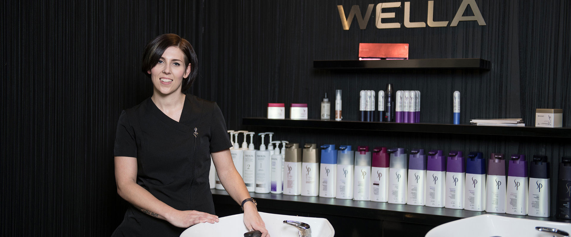 Hairdressing student smiling while they stand next to the shampoo section of the salon, with chair basins and products next to them.