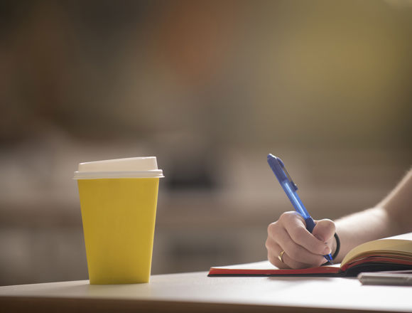 Disposable yellow coffee cup on a desk while a student is taking notes in a notebook.