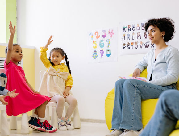 Group of young children holding up their hands in a nursery, with their teacher sitting near them.