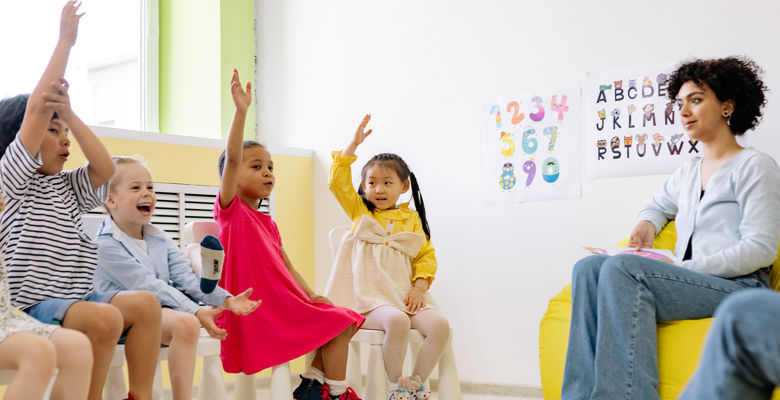 Group of young children holding up their hands in a nursery, with their teacher sitting near them.