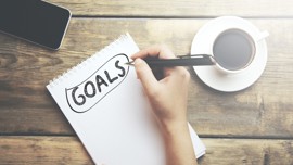 Notebook on a table surrounded by a mobile phone and cup of coffee. The word "Goals" is written on the notebook page.