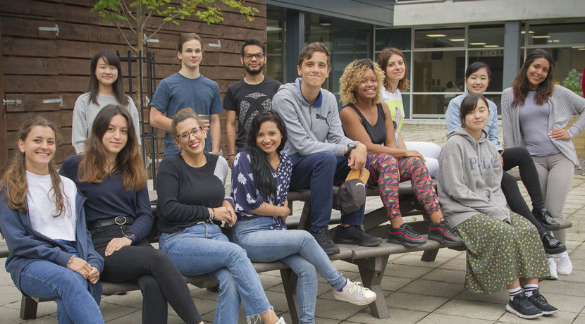 Group of students sitting on a bench, posing for a photo outside a college campus building.