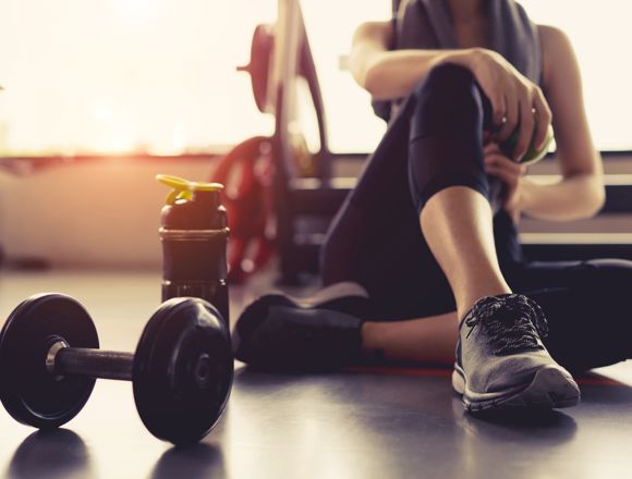 Person in fitness clothing sitting down in the gym with a water bottle and weights to their side.