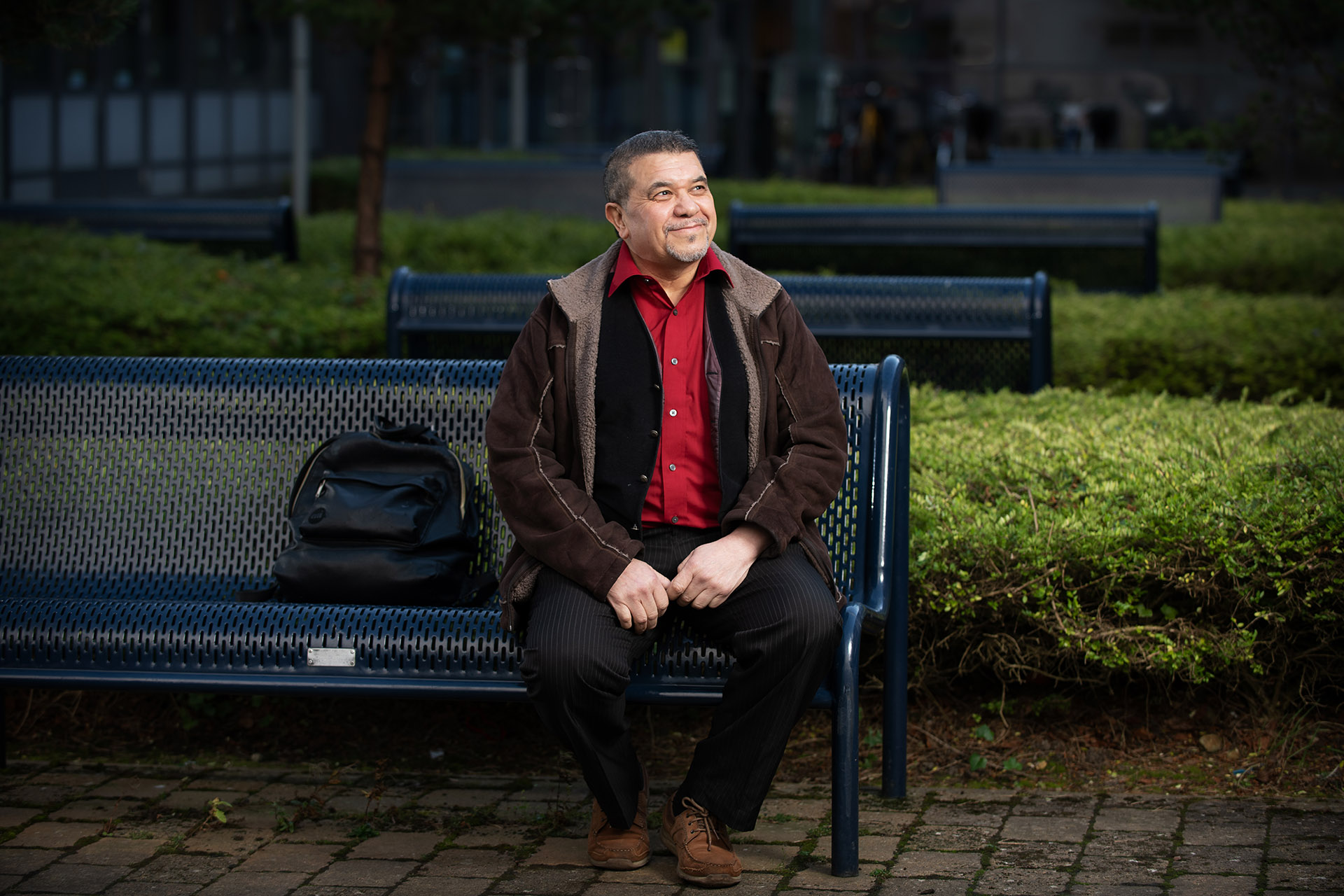 A male sitting on a blue metal bench outside a campus building with their backpack next to them.