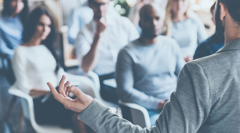 Person in a suit presenting in front of a group of people who are sitting down.