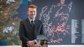 Young student wearing a suit, holding a Historic Scotland magazine at the front of a classroom.