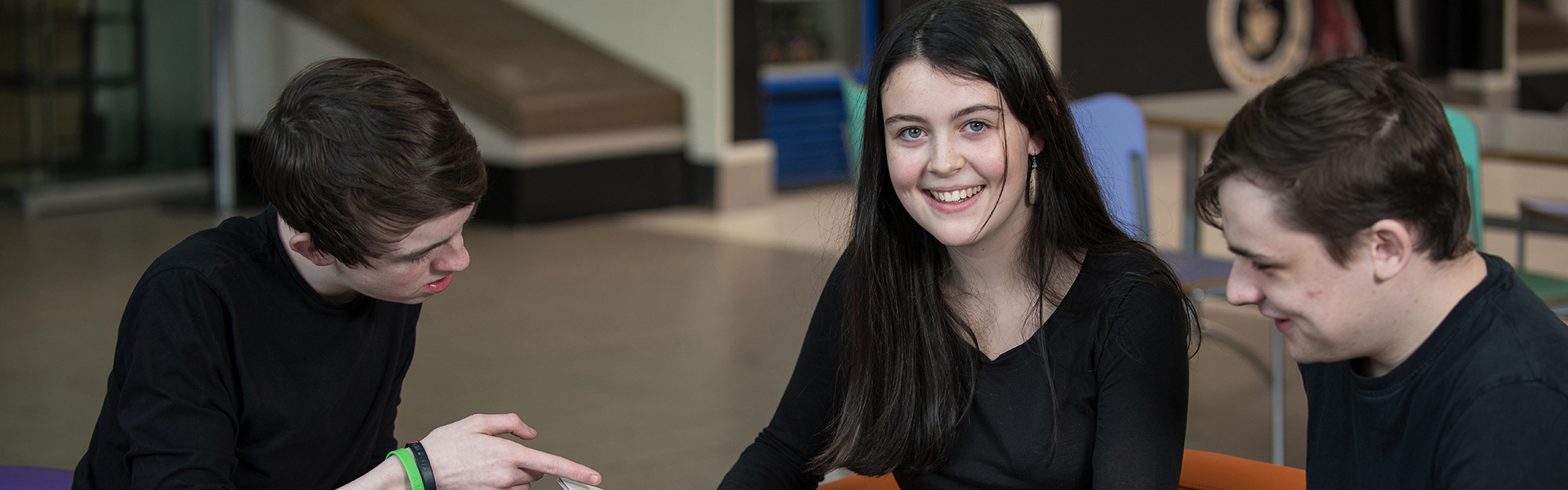 Two males looking at a book together with a female sitting in the middle looking directly at the camera and smiling.