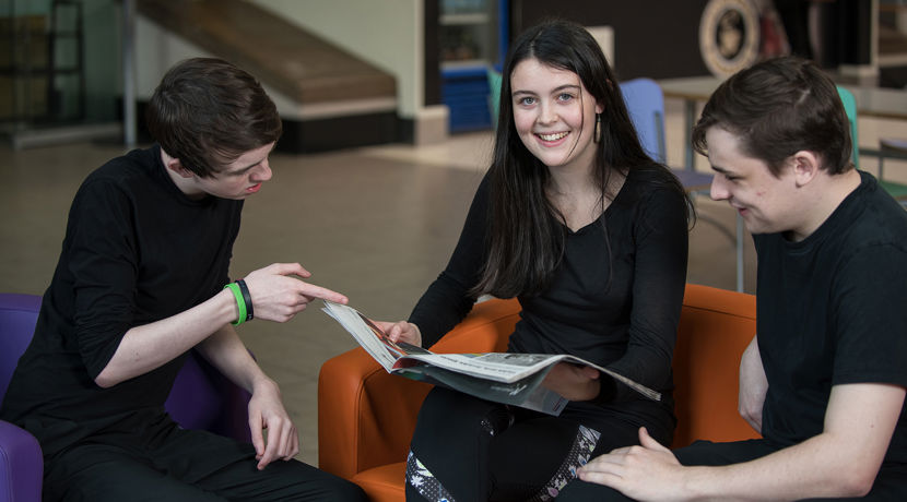 Two males looking at a book together with a female sitting in the middle looking directly at the camera and smiling.