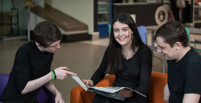 Two males looking at a book together with a female sitting in the middle looking directly at the camera and smiling.