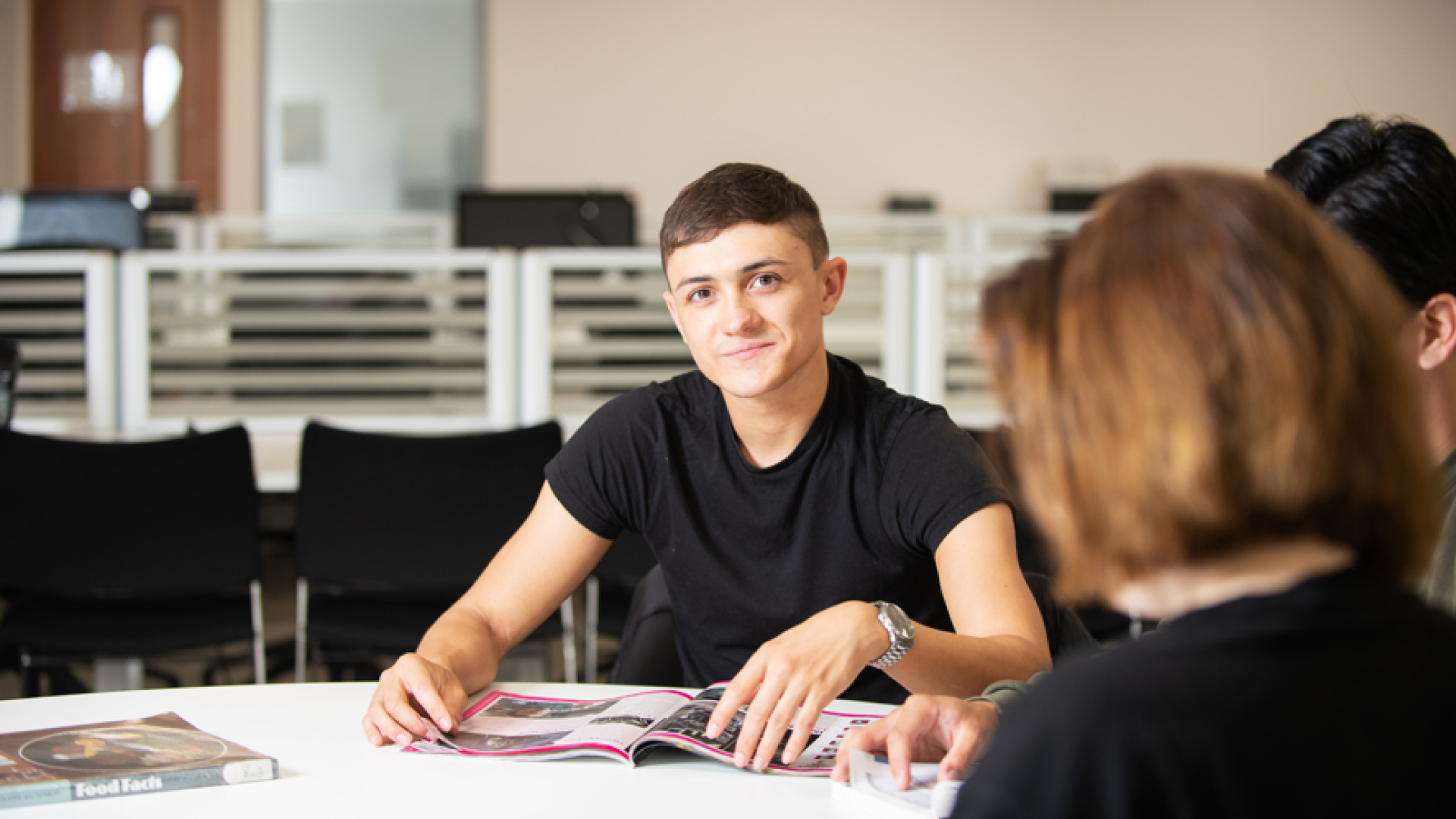 A man looks directly at the camera smiling, he has an open brochure on the table in front of him.