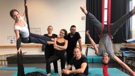 A group of six people in a dance studio, showcasing various acrobatic poses with colorful mats and equipment in the background.