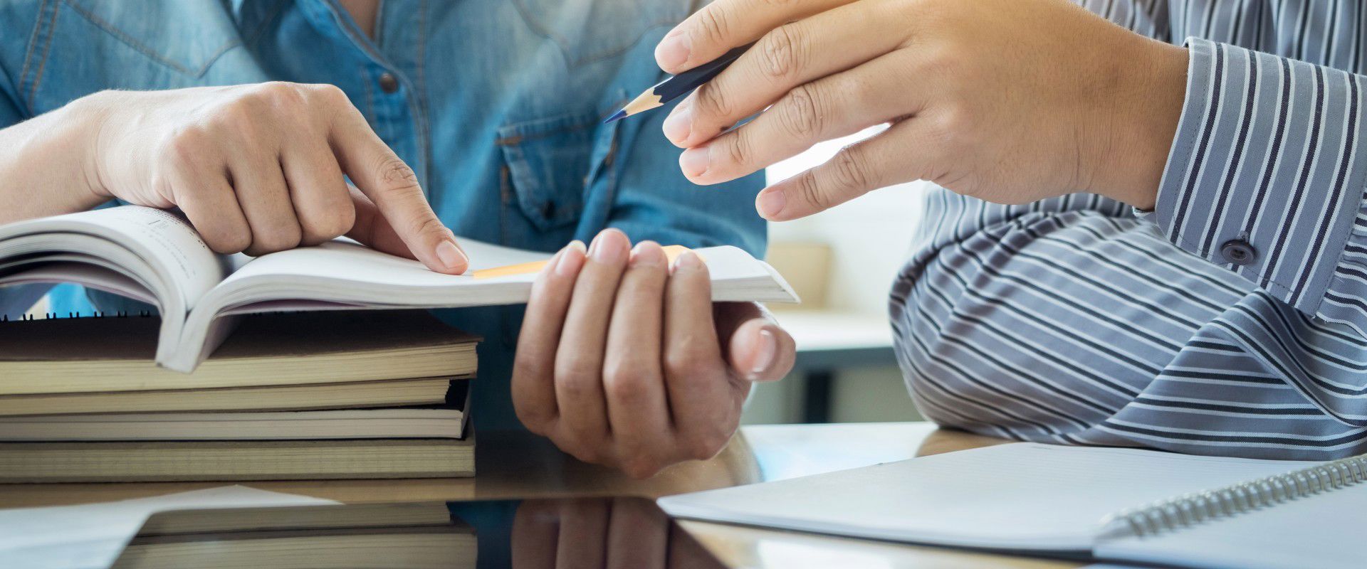 Two students are pointing at something in an open textbook, on top of a stack of closed textbooks.