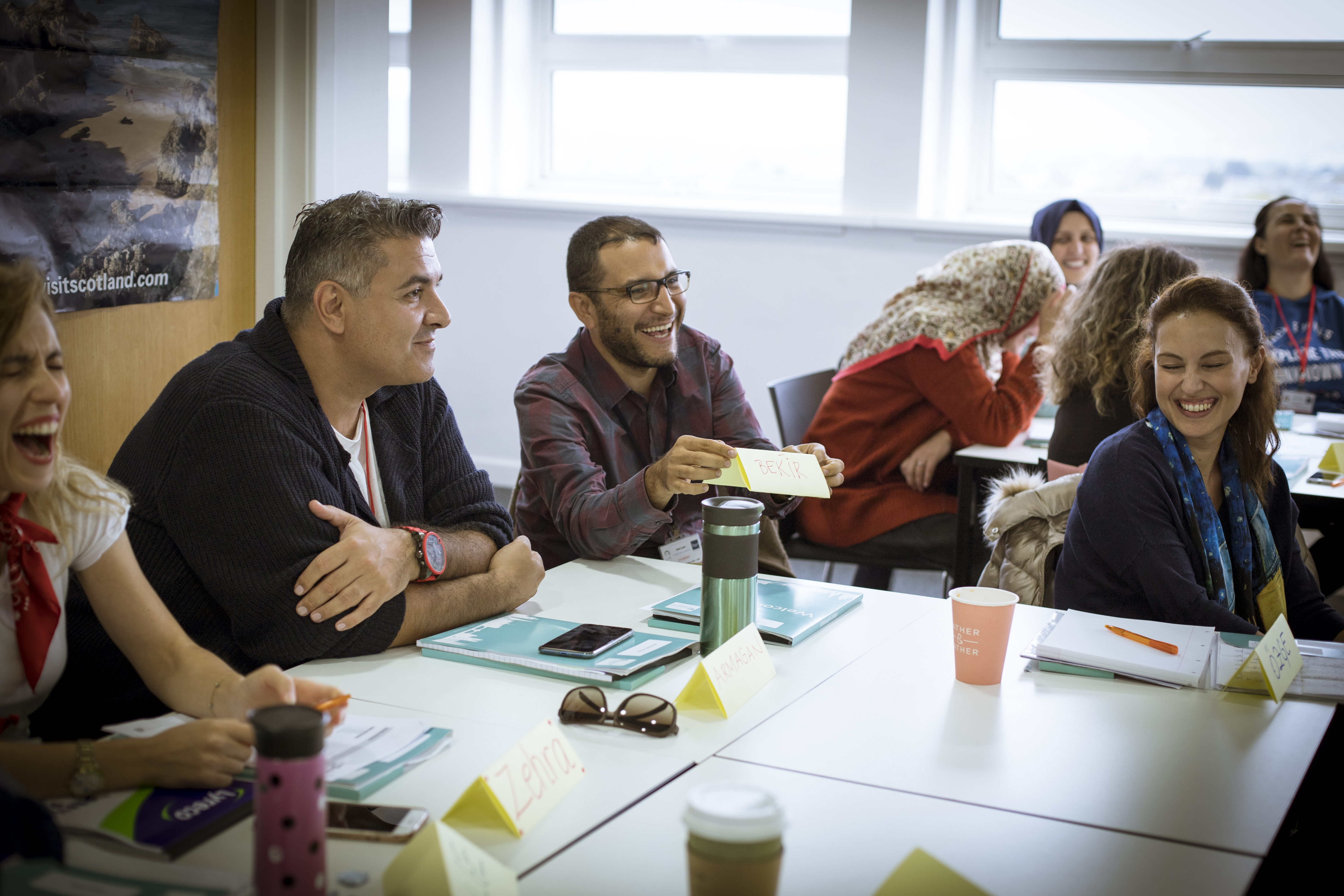 A group of people sit around a conference table, smiling and laughing, holding materials. Name tags and notebooks are visible on the table.