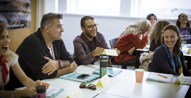 A group of people sit around a conference table, smiling and laughing, holding materials. Name tags and notebooks are visible on the table.