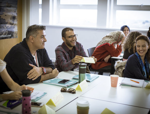 A group of people sit around a conference table, smiling and laughing, holding materials. Name tags and notebooks are visible on the table.