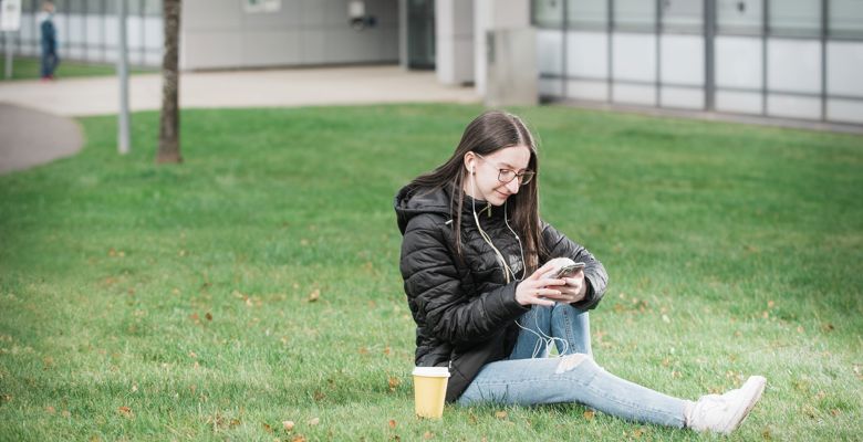 A young student is sitting on the grass looking at their phone at Granton campus. 